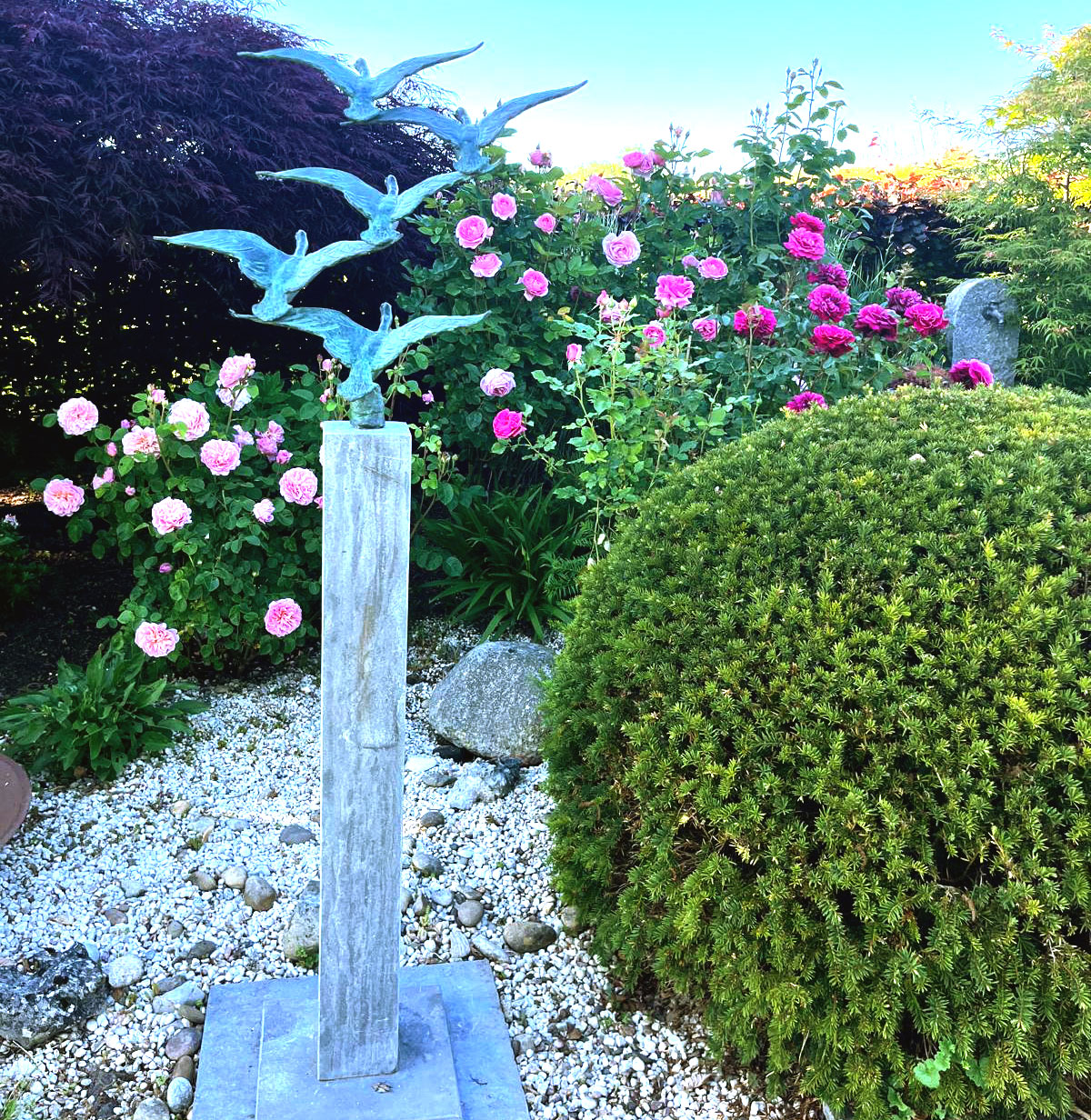 Kugelförmig geschnittene Sträucher mit einer Stele aus Granit, die fliegende Vögel darstellt, dahinter Rosensträucher